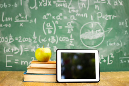 An iPad next to stacked books with an apple on top all in front of a blackboard with complex formulas.